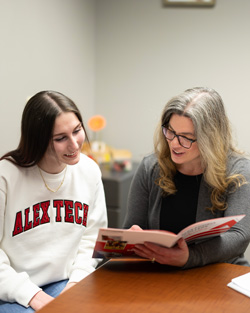 A middle aged woman holds a pamphlet and shows it to a young woman wearing an Alex Tech shirt