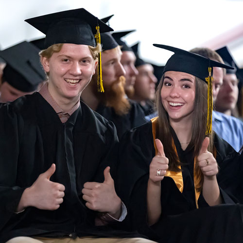 Two students in graduation cap and gown looking at the camera, both giving two thumbs up.