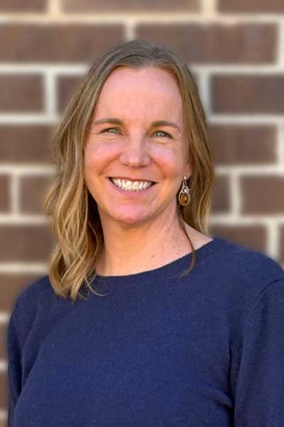 Kerri Barnstuble_Saved-for-Web Headshot of woman wearing navy blue sweater and standing against a brown brick wall