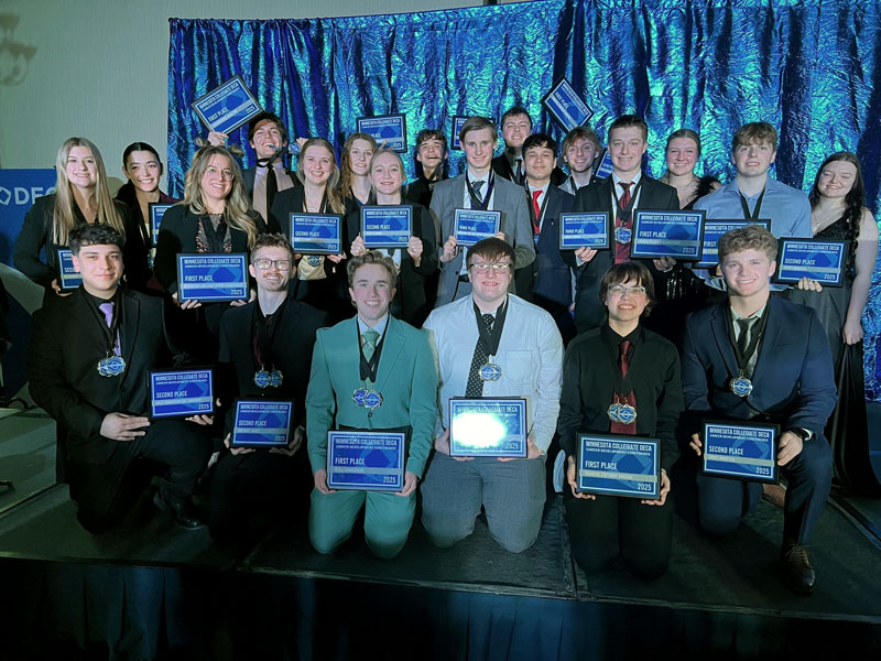MN Collegiate DECA Awards 2025. Students stand in front of a blue backdrop wearing medals and holding award plaques.