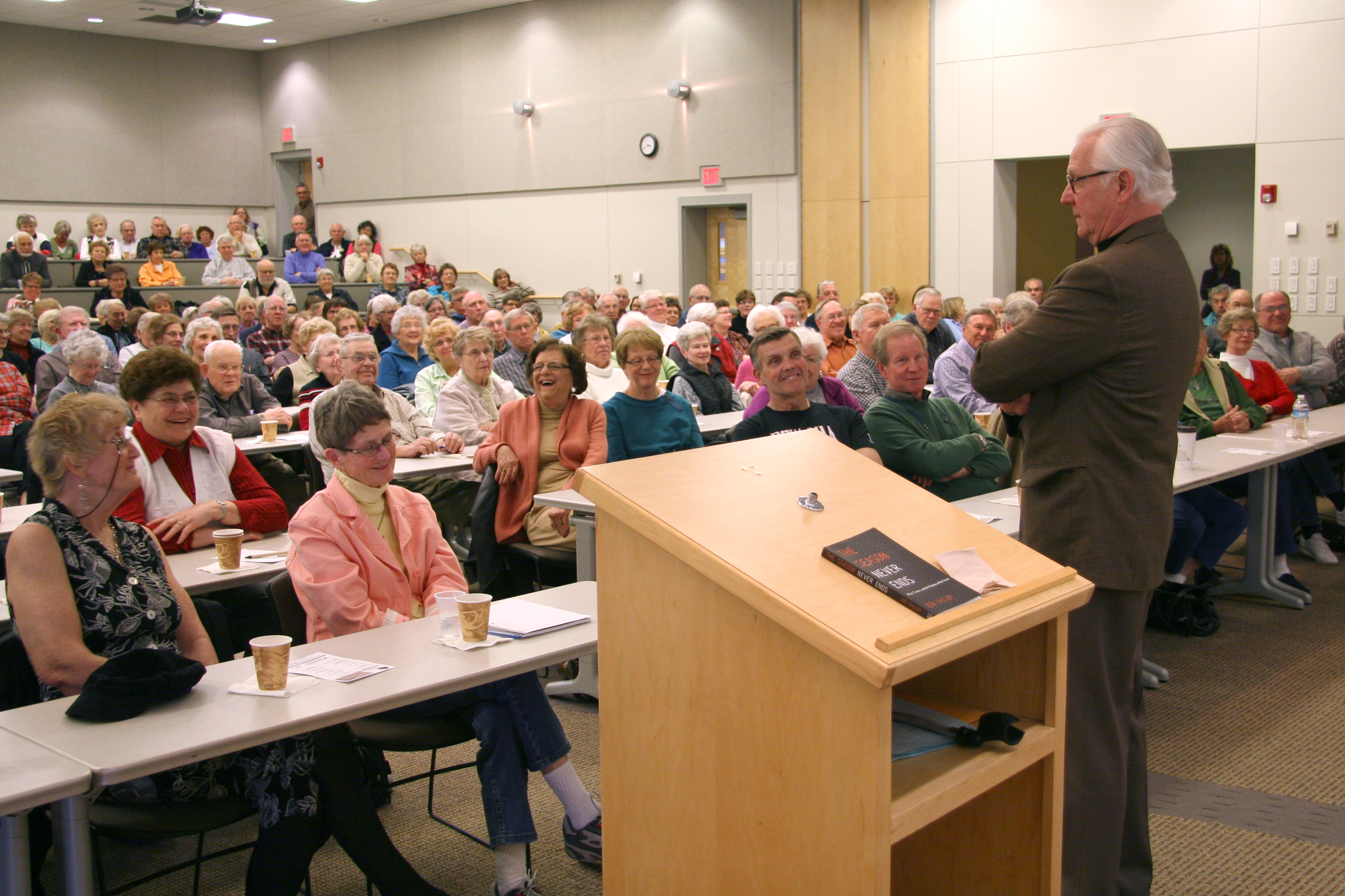 A male speaker in a brown business suit stands by a podium and addresses a large room full of senior citizen attendees seated at tables